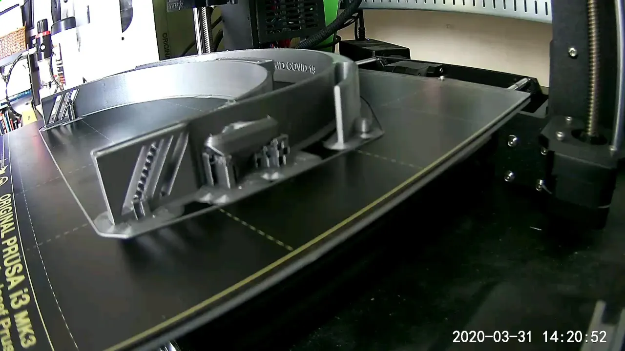 A Prusa i3 MK3 3D printer mid-print, building gray face-shield headbands labeled MD COVID 19 on its print bed
